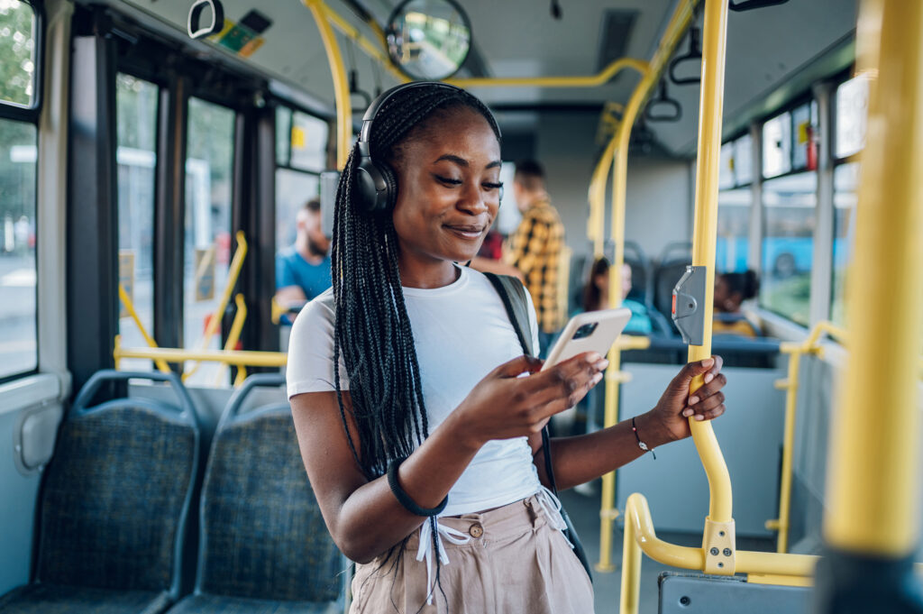 person standing in a transit bus with headphones on, smiling at phone.