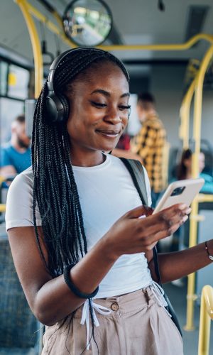 person standing in a transit bus with headphones on, smiling at phone.